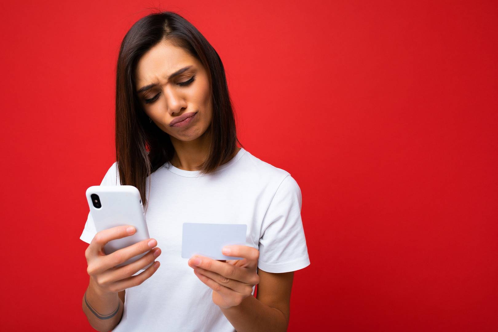 Kobieta w białej koszulce trzyma w dłoni smartfona i zdrapkę z kodem na doładowanie telefonu na kartę. Jest skoncentrowana i wpatruje się w cyfry na kuponie.