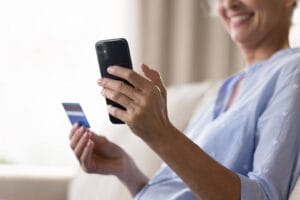 Starsza kobieta trzyma w jednej dłoni telefon, a w drugiej – kartę z kodem na doładowanie konta. Wpisuje 14-cyfrowy kod, by zasilić środki na karcie.