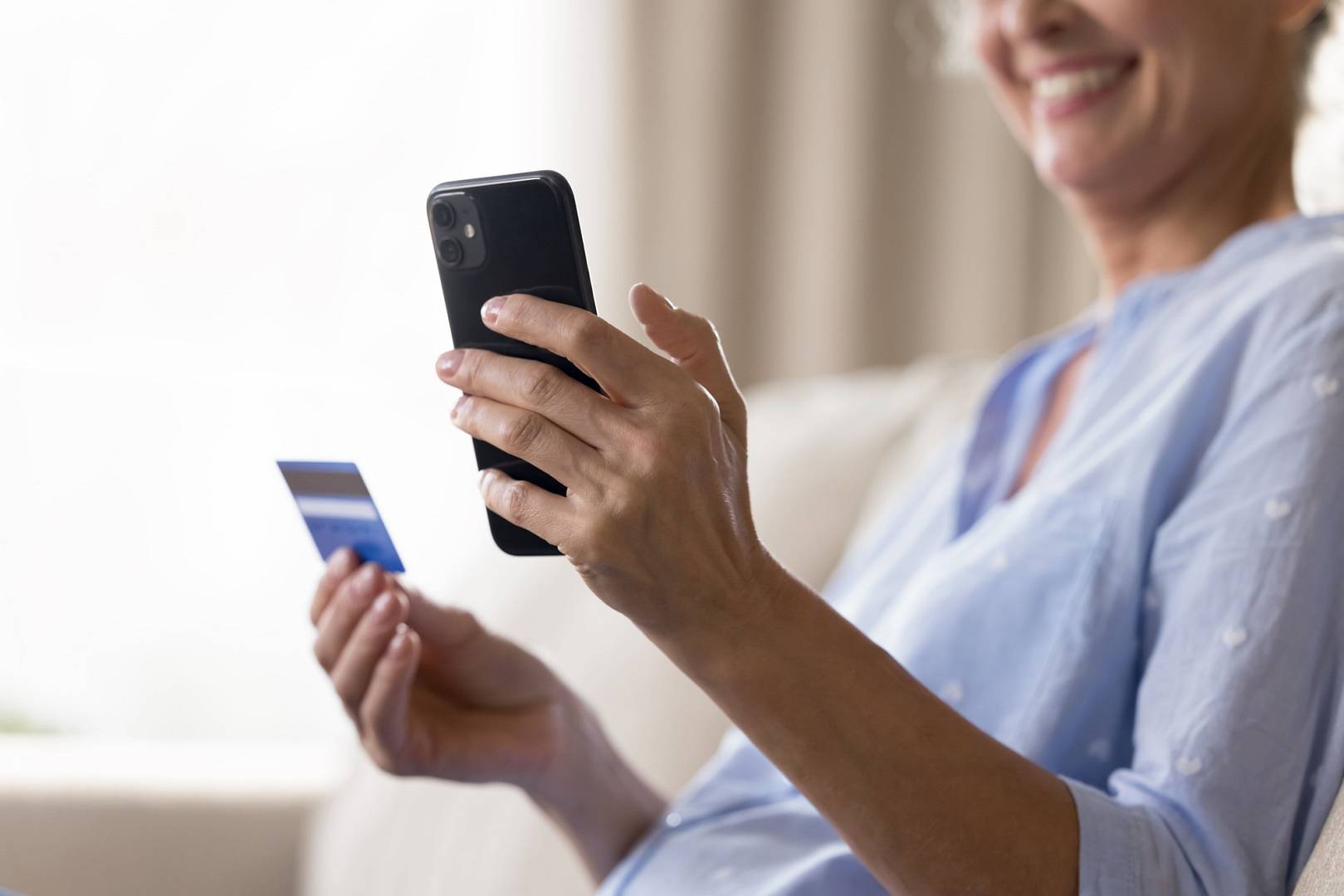 Starsza kobieta trzyma w jednej dłoni telefon, a w drugiej – kartę z kodem na doładowanie konta. Wpisuje 14-cyfrowy kod, by zasilić środki na karcie.