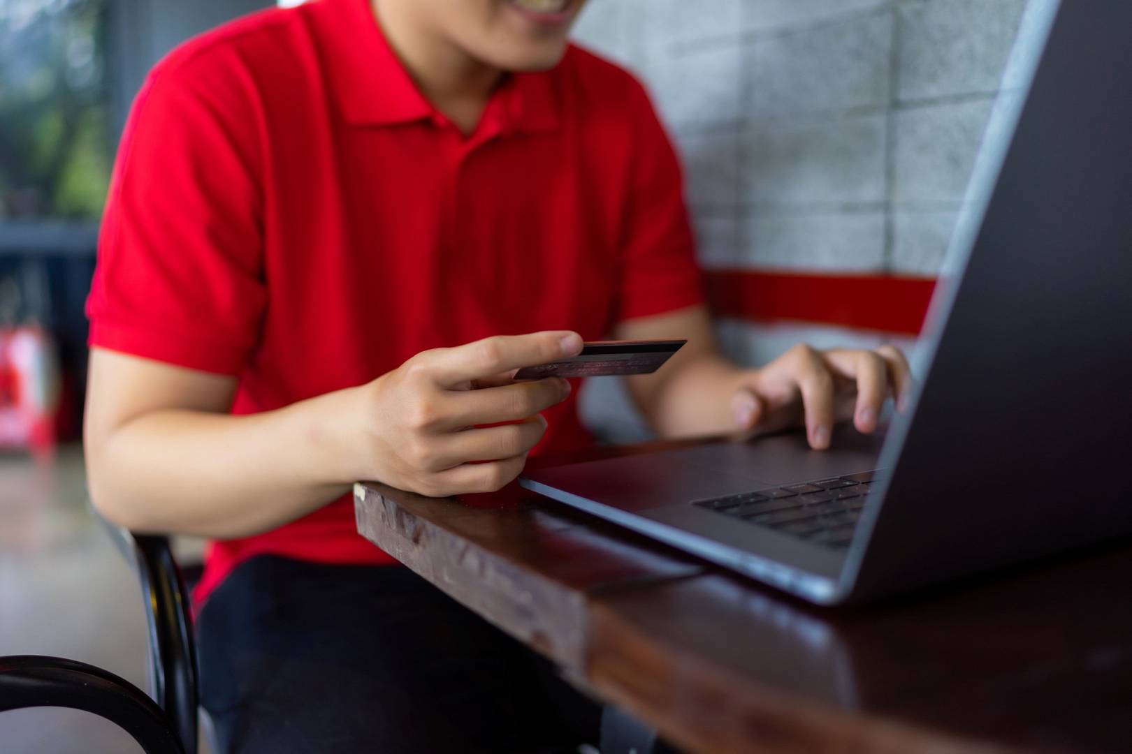 Mężczyzna w czerwonej koszuli kupuje przez internet doładowanie telefonu na kartę. Korzysta w tym celu z laptopa, gdy siedzi przy stoliku w kawiarni.